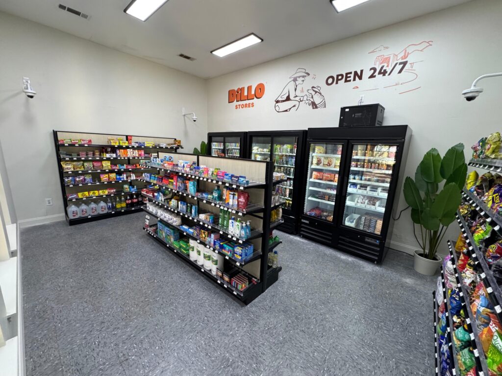 Small convenience store with shelves of snacks, drinks, and groceries. Refrigerators line the back wall. A mural with Dillo Stores and OPEN 24/7 is on the wall above the fridges. The floor is gray tile.