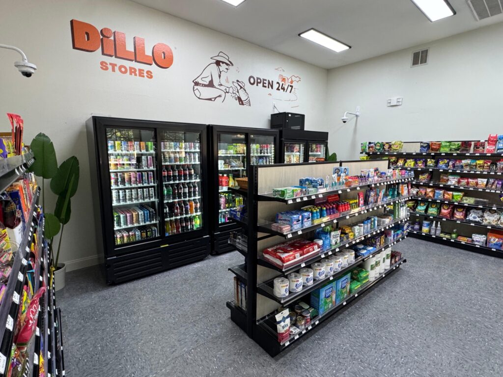 A small convenience store with shelves of snacks, drinks, and household items. Refrigerators filled with beverages line one wall. A mural reads Dillo Stores Open 24/7 with an armadillo illustration above.