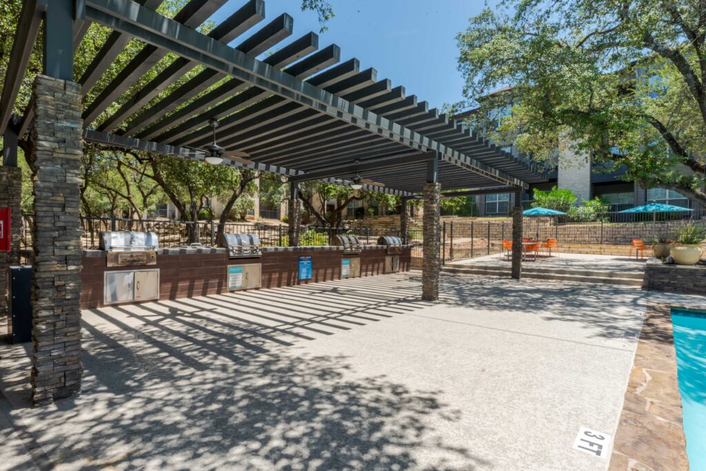 Outdoor patio area with a pergola, stone pillars, and a built-in grill station beside a swimming pool. There are trees, lounge seating, and tables with umbrellas in the background under a clear sky.