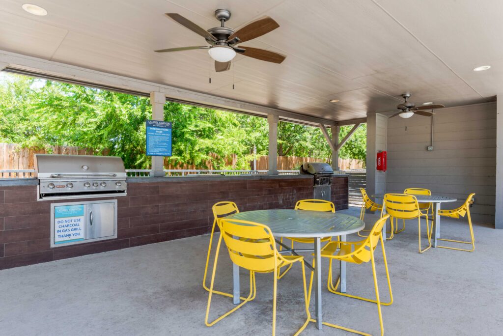 Covered outdoor patio with two metal tables and bright yellow chairs, ceiling fans, a stainless steel barbecue grill, and trees visible beyond a wooden fence in the background.