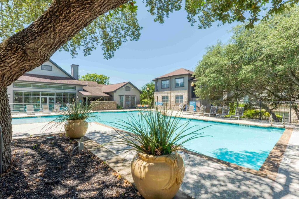 A sunny outdoor swimming pool surrounded by lounge chairs and trees, with large ceramic planters in the foreground and residential buildings in the background.