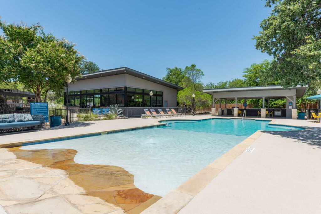 Outdoor swimming pool with shallow entry, surrounded by trees, lounge chairs, and shaded seating areas. Modern building and covered patio visible in the background under a clear blue sky.