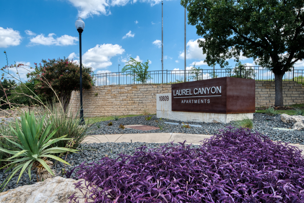 A landscaped entrance with purple and green plants surrounds a stone sign reading Laurel Canyon Apartments 10009 under a sunny sky with scattered clouds, showcasing one of the most inviting San Antonio Apartments.