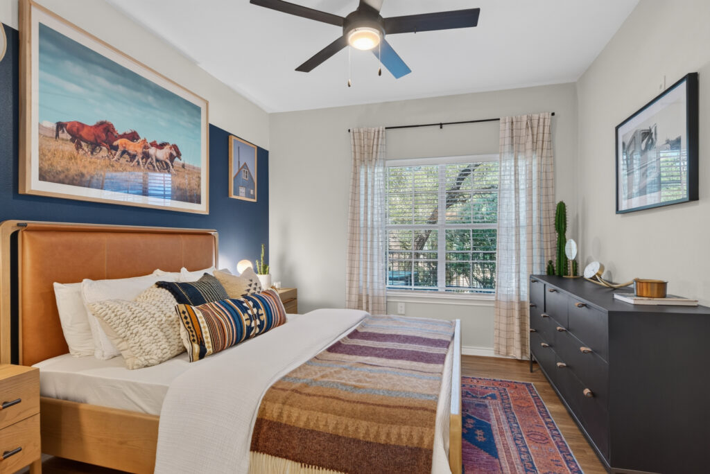 A cozy bedroom in San Antonio Apartments features a leather headboard, colorful pillows, a blanket, a dresser, large window with sheer curtains, ceiling fan, and wall art showcasing horses and a barn.