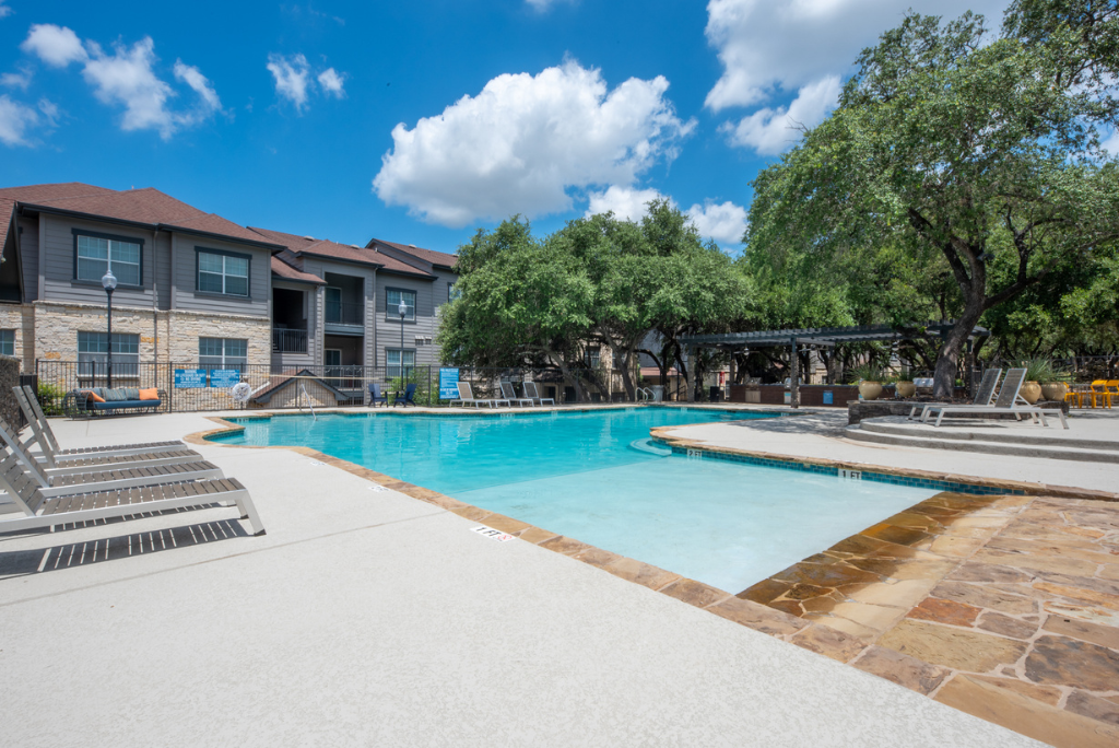 Outdoor swimming pool with lounge chairs, surrounded by trees and apartment buildings under a blue sky with scattered clouds—experience the relaxed lifestyle of San Antonio Apartments.