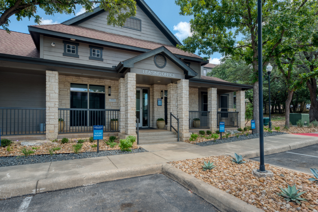 A modern leasing office building for San Antonio Apartments features a stone and wood exterior, a covered entryway, and a ramp for accessibility. Signs and landscaped areas with small plants are visible in front.