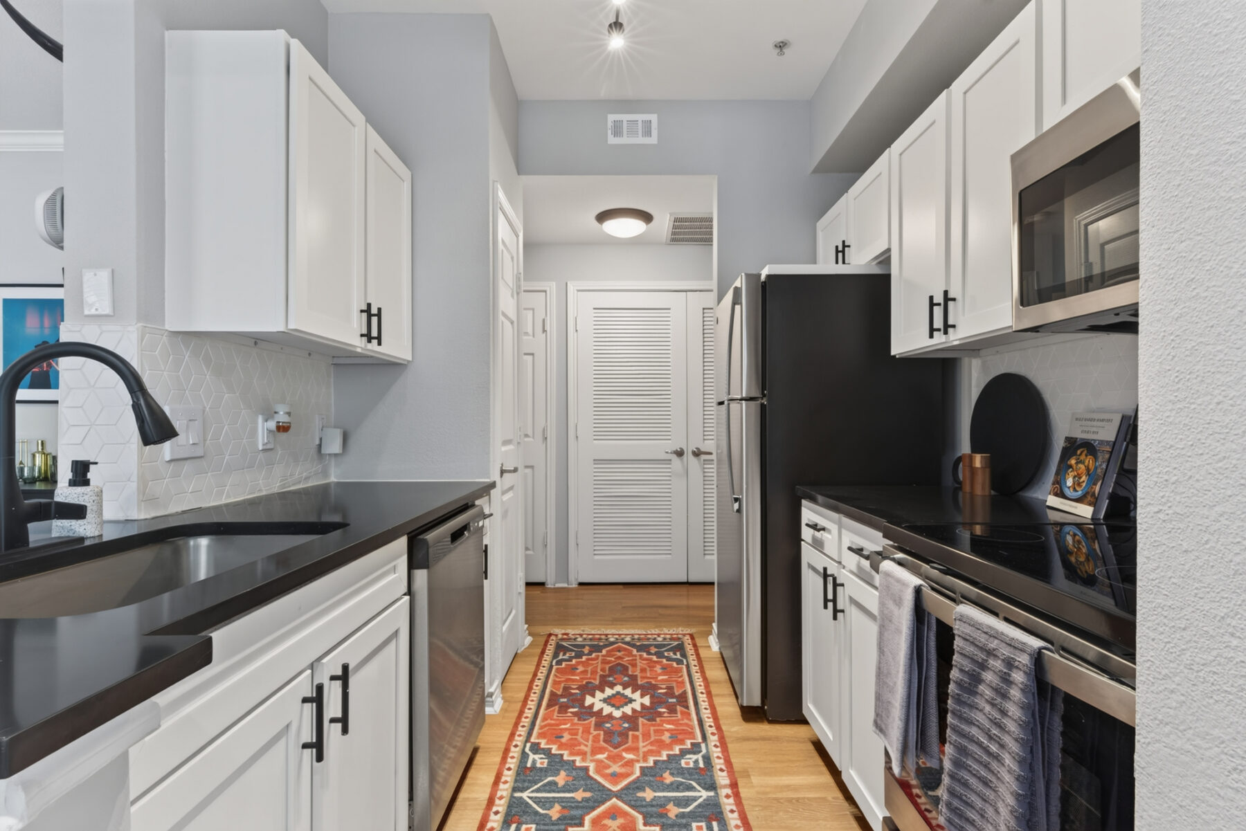Modern kitchen with white cabinets, black countertops, stainless steel appliances, and a geometric-patterned rug on a wooden floor. A hexagon tile backsplash is visible, with towels hanging on the oven door—an inviting space that perfectly blends style and functionality, commonly found in upscale San Antonio Apartments.
