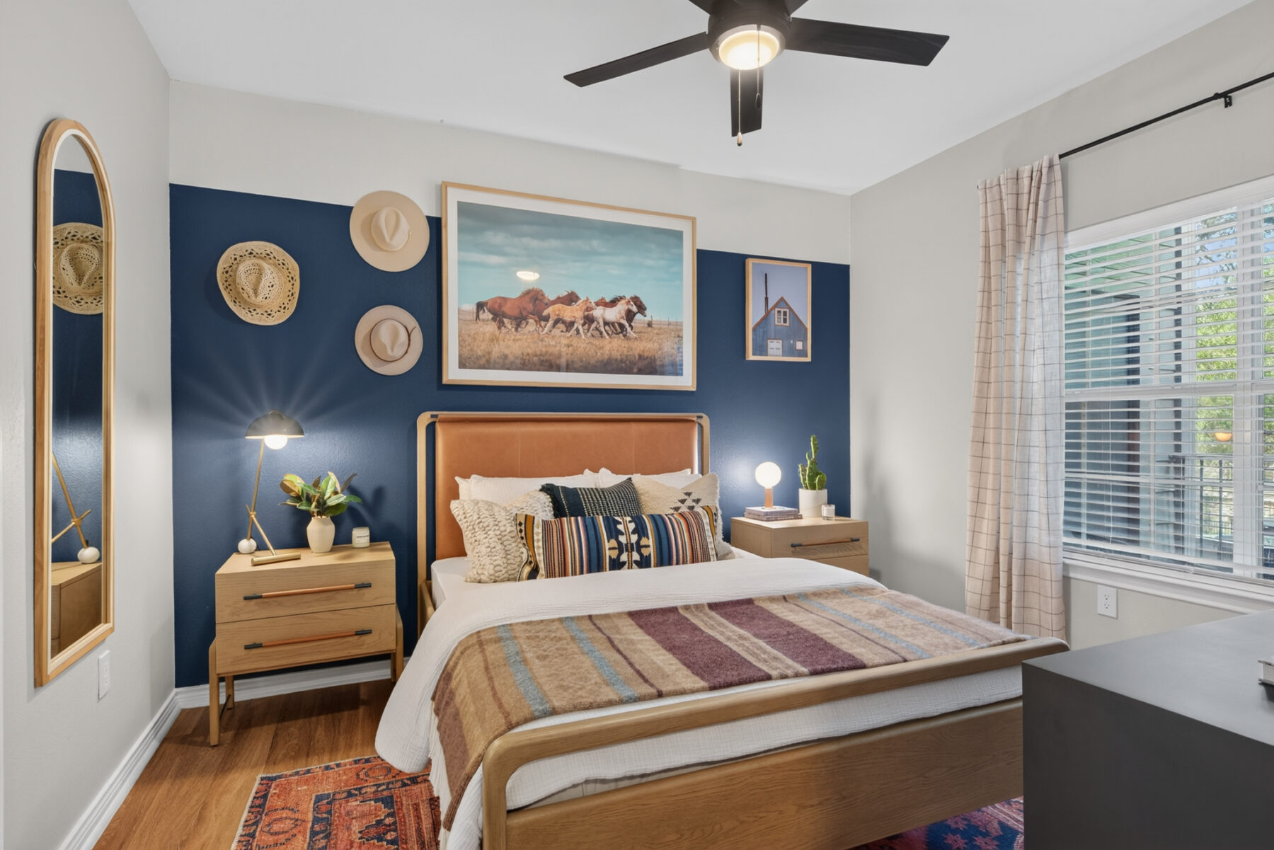 A cozy bedroom with a blue accent wall, wooden bed and nightstands, a ceiling fan, western decor, a horse photo above the bed, a large window with blinds, and various pillows and plants.