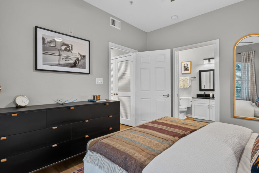 A modern bedroom with a striped blanket on the bed, a black dresser, a framed photo on the wall, and an open door showing a bathroom with a sink, mirror, and toilet. Natural light comes in from a window.