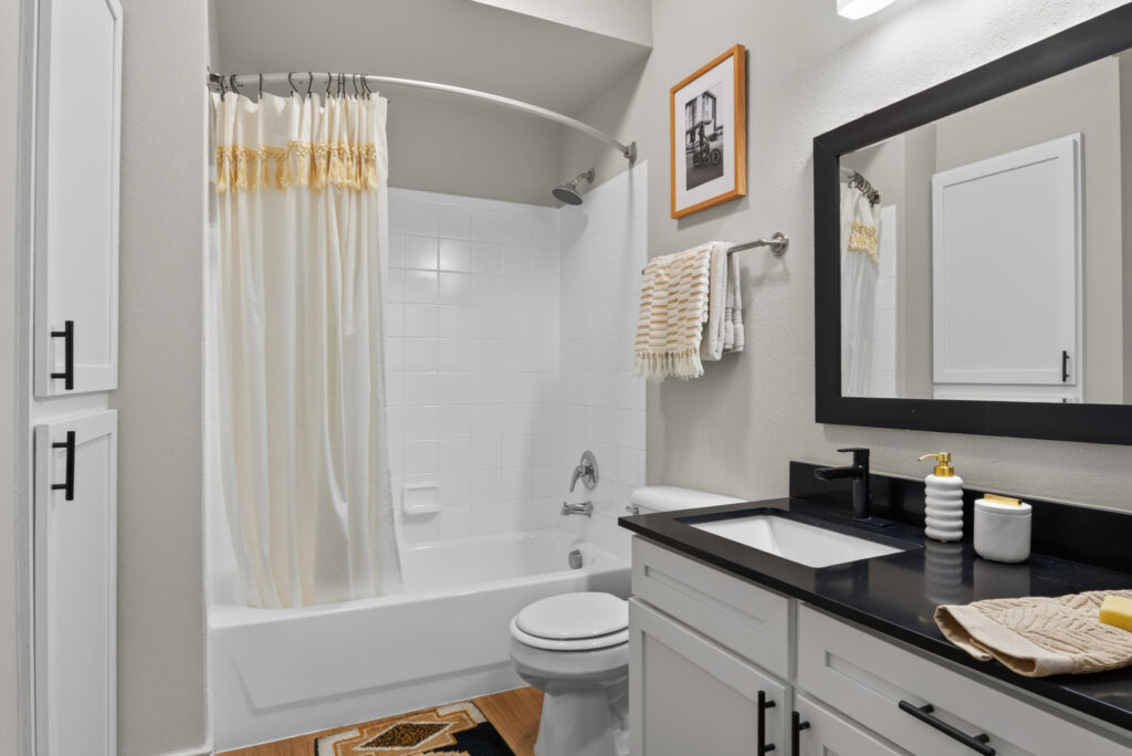 Modern bathroom with white cabinets, black countertop, bathtub with cream shower curtain, toilet, mirror, and decorative items like towels, soap dispensers, and framed art on a light-colored wall.